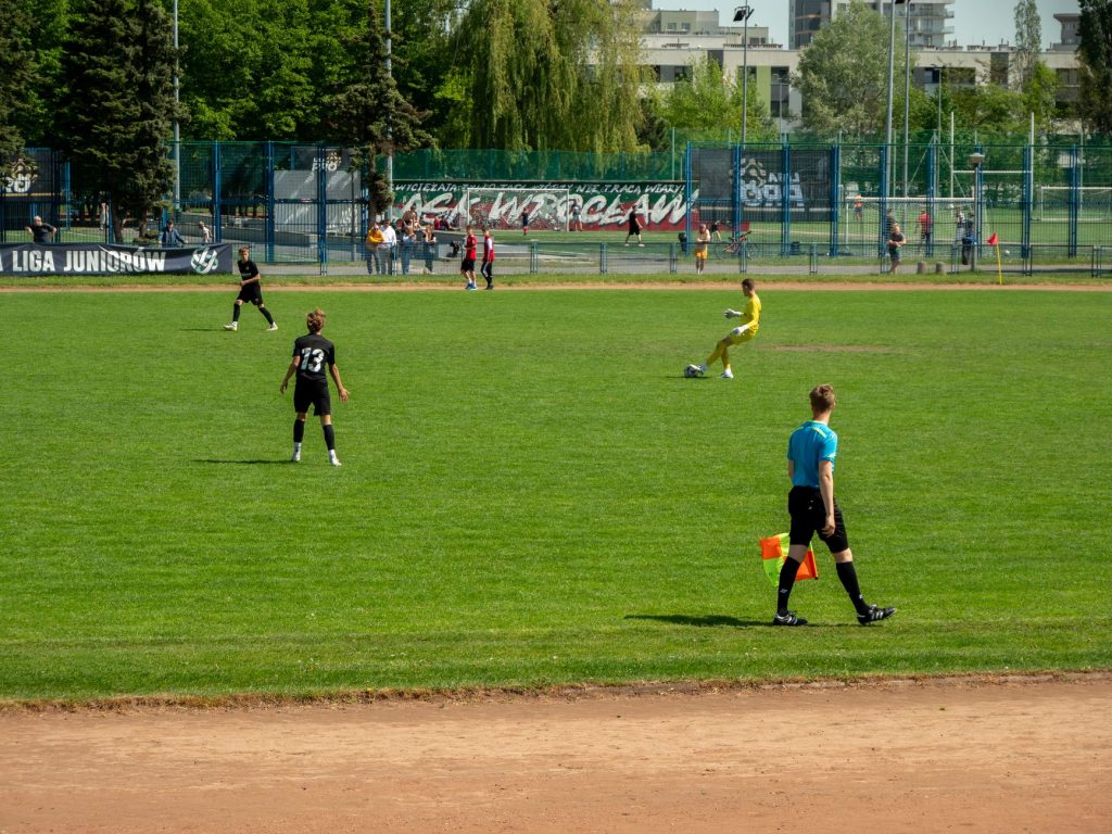 Youth soccer game at Wrocław sports field on a sunny day with players and referee in action.